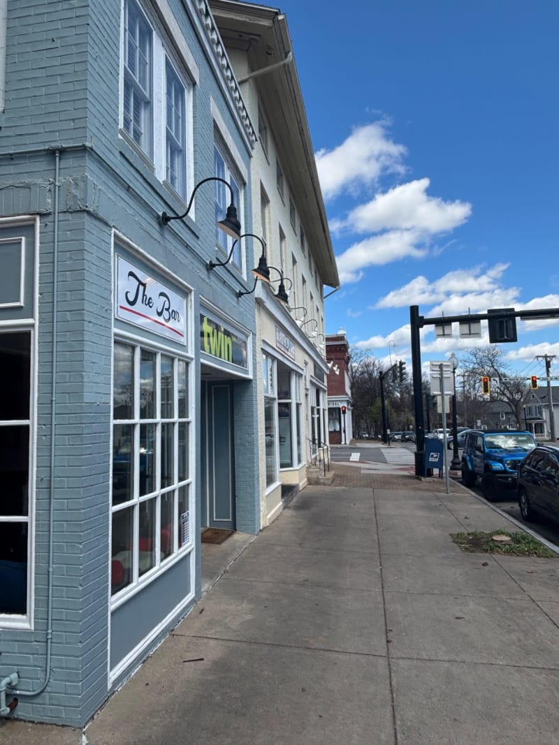 # Alt Text
Sidewalk view of "The Bar" storefront on a sunny day with clear blue sky and local buildings in the background.