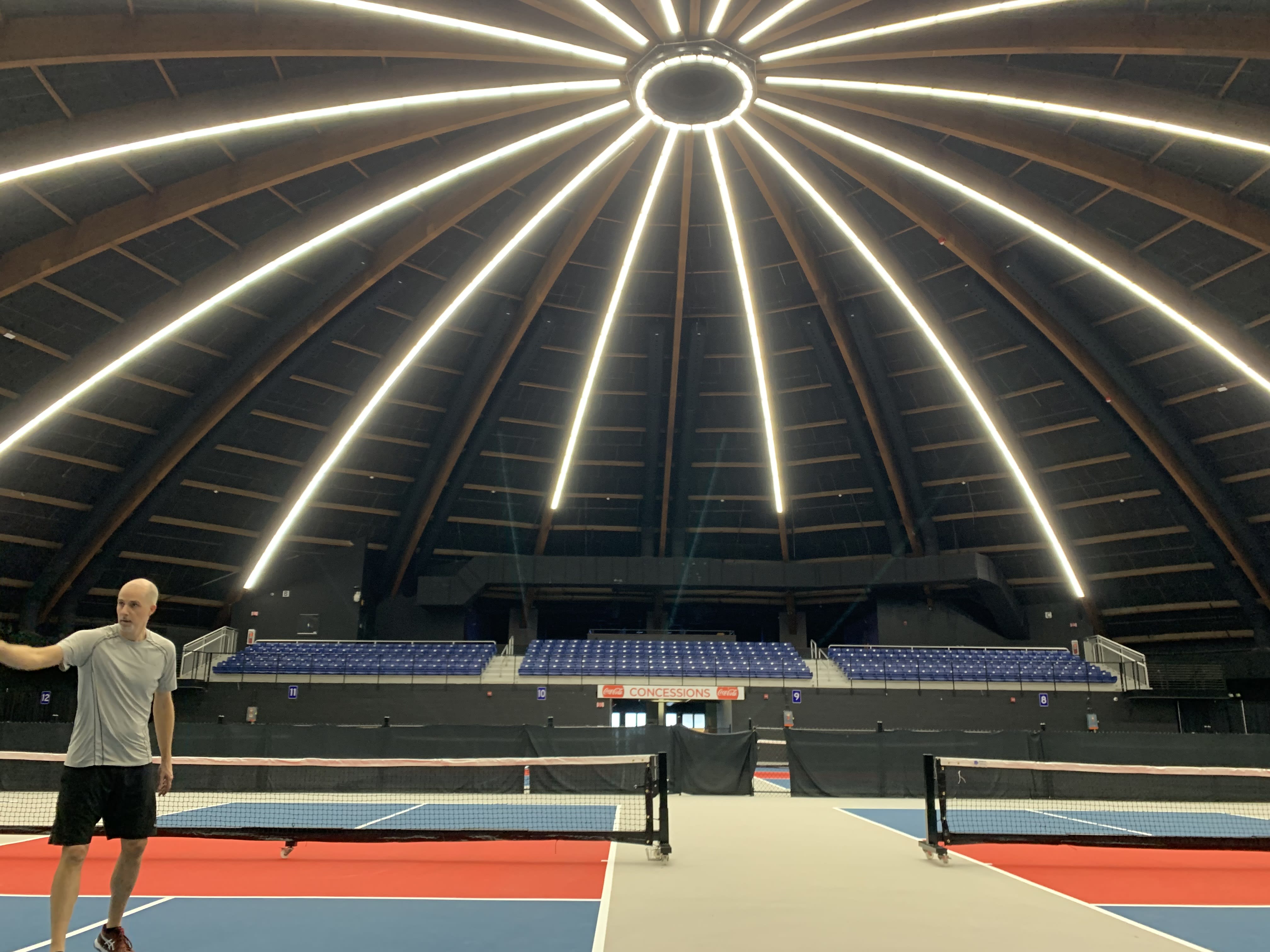 # Alt Text
Man standing on indoor tennis court under distinctive domed ceiling with radiating LED lighting strips.