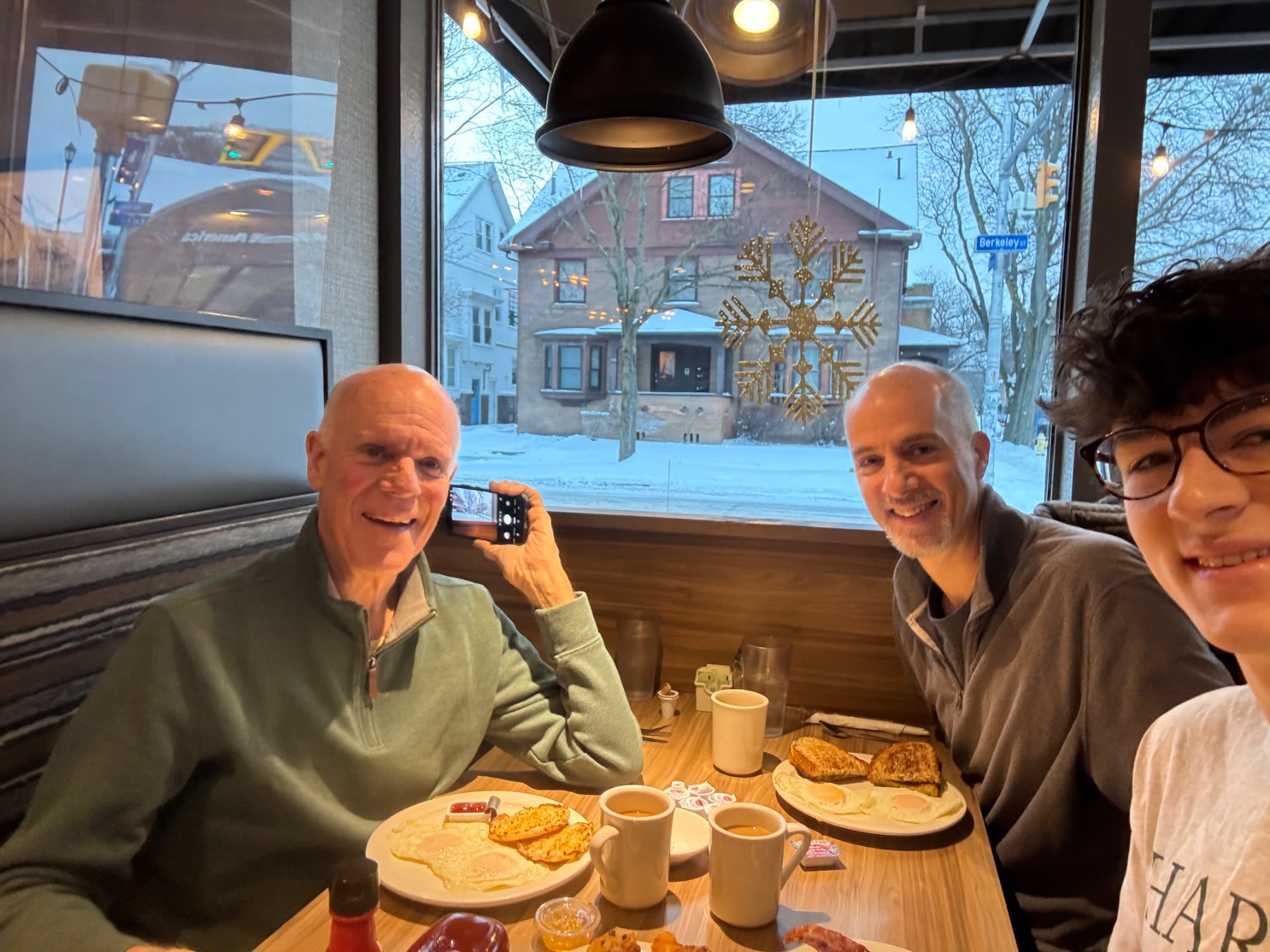 Three people enjoy breakfast together at a snowy winter café with decorated buildings visible through the window.