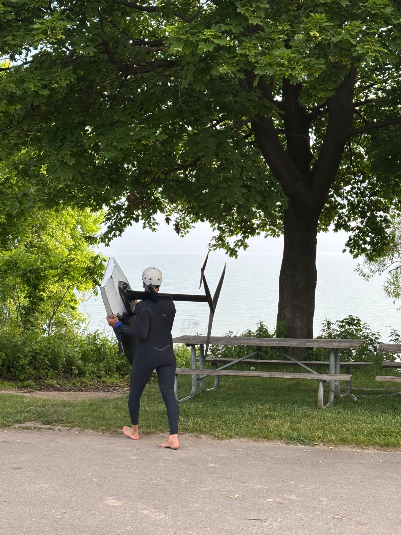 A kiteboarder in a wetsuit carries their board and kite near a waterfront picnic area under large trees.