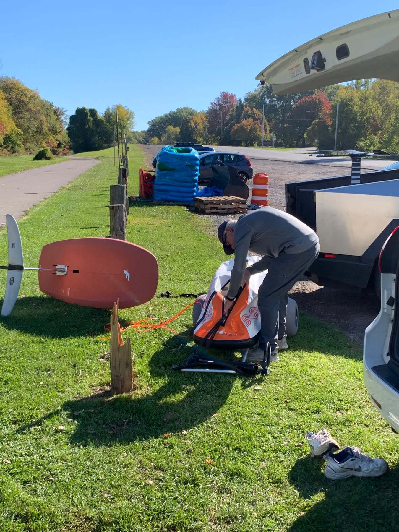 A person inspects an orange and white kiteboarding board at a grass landing area near a runway and parked vehicles.