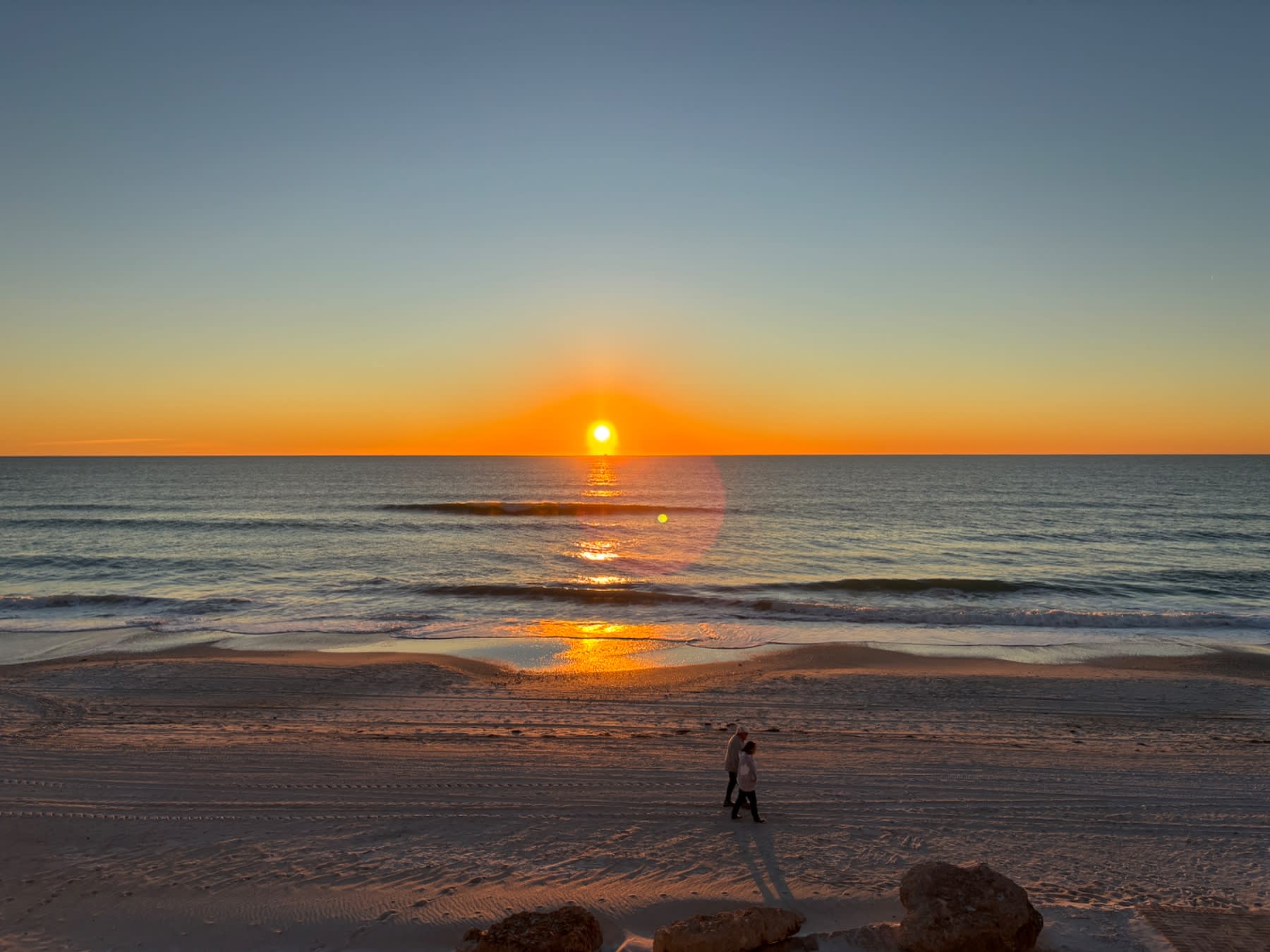 A surfer walks along the beach at sunrise with golden sunlight reflecting off the ocean waves.