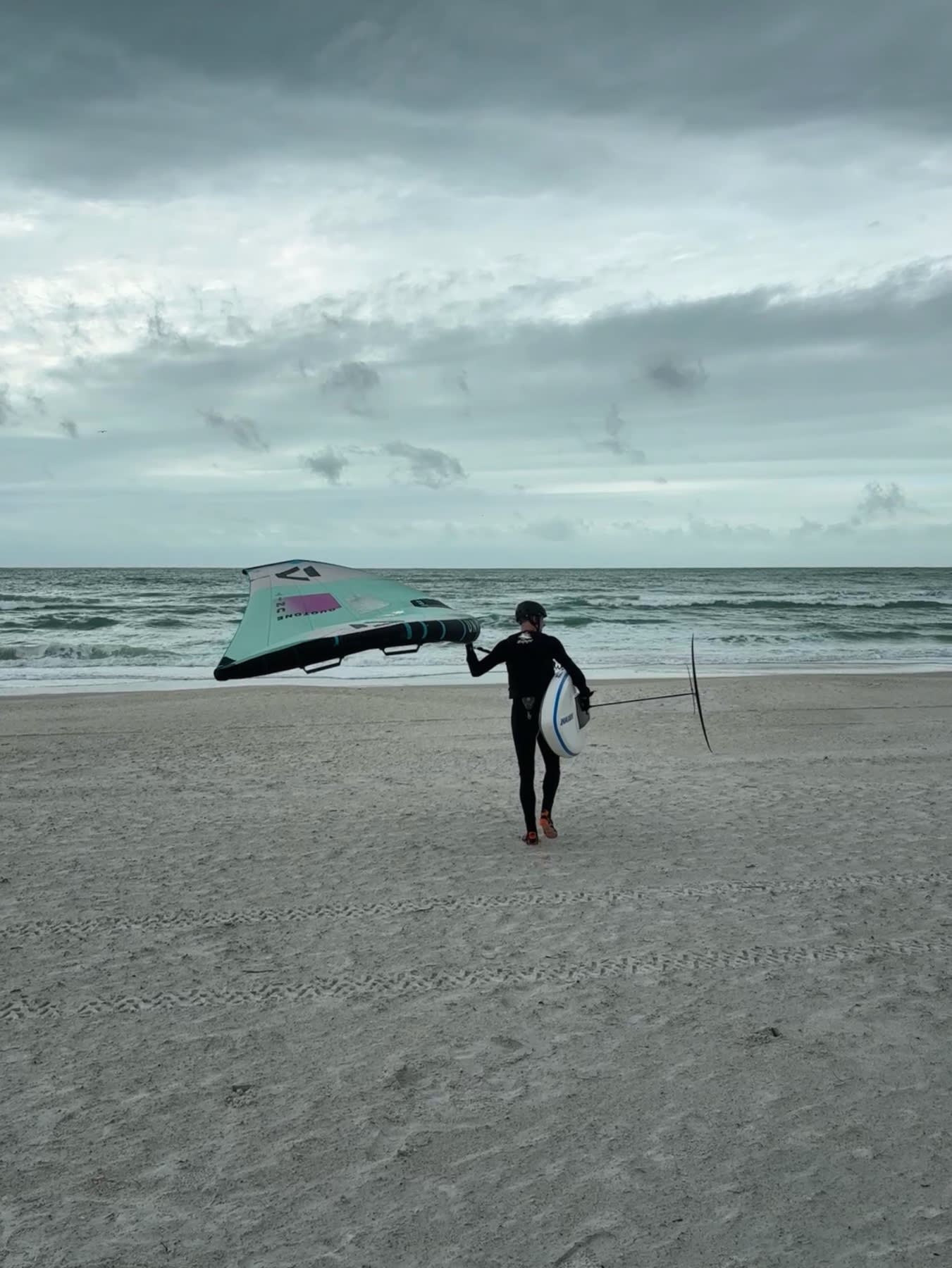 A kite surfer in a wetsuit carries their kite and board toward the ocean on a sandy beach under cloudy skies.