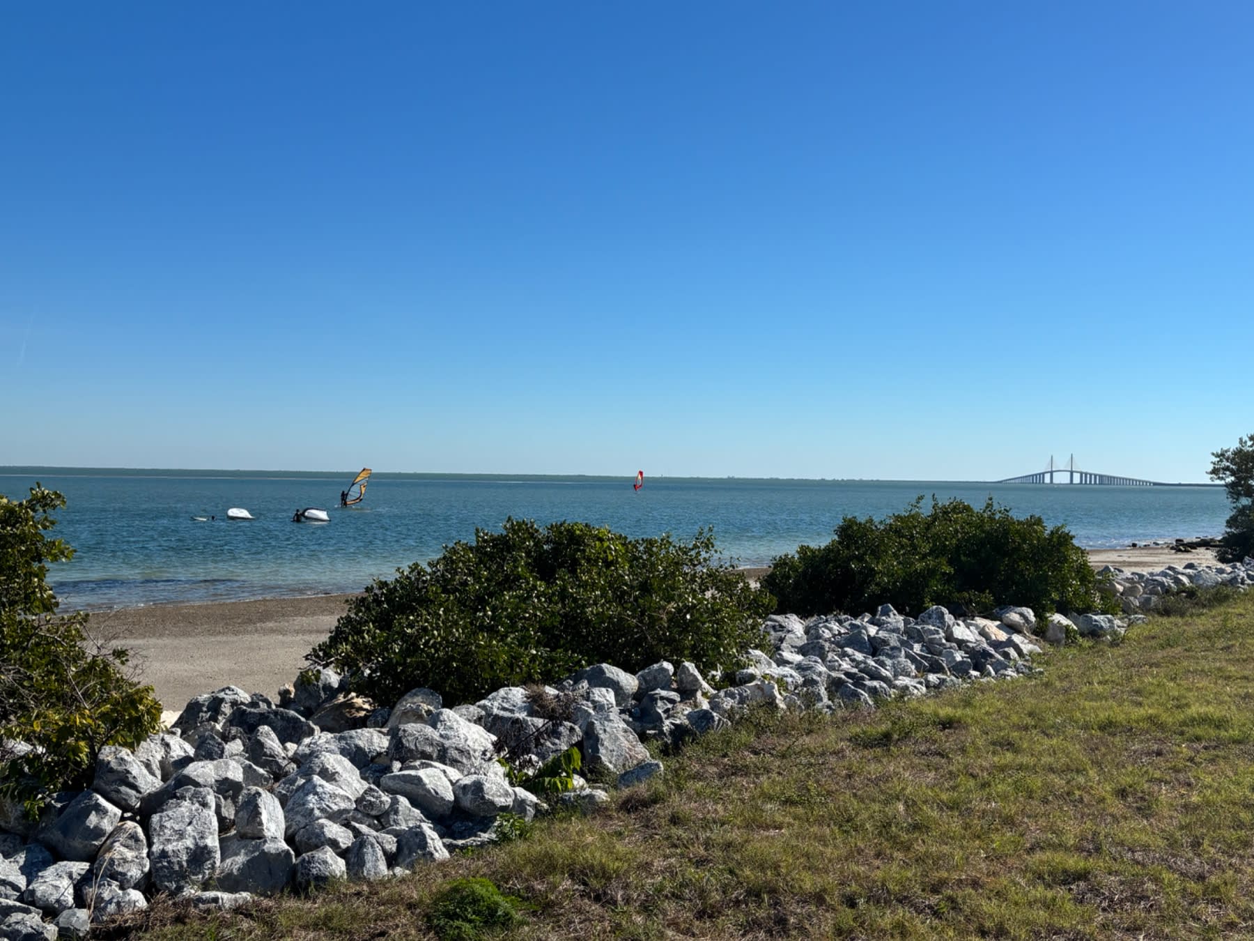 "Beach cove with rocky shoreline, calm blue waters, sailboats, and a suspension bridge visible in the distance."