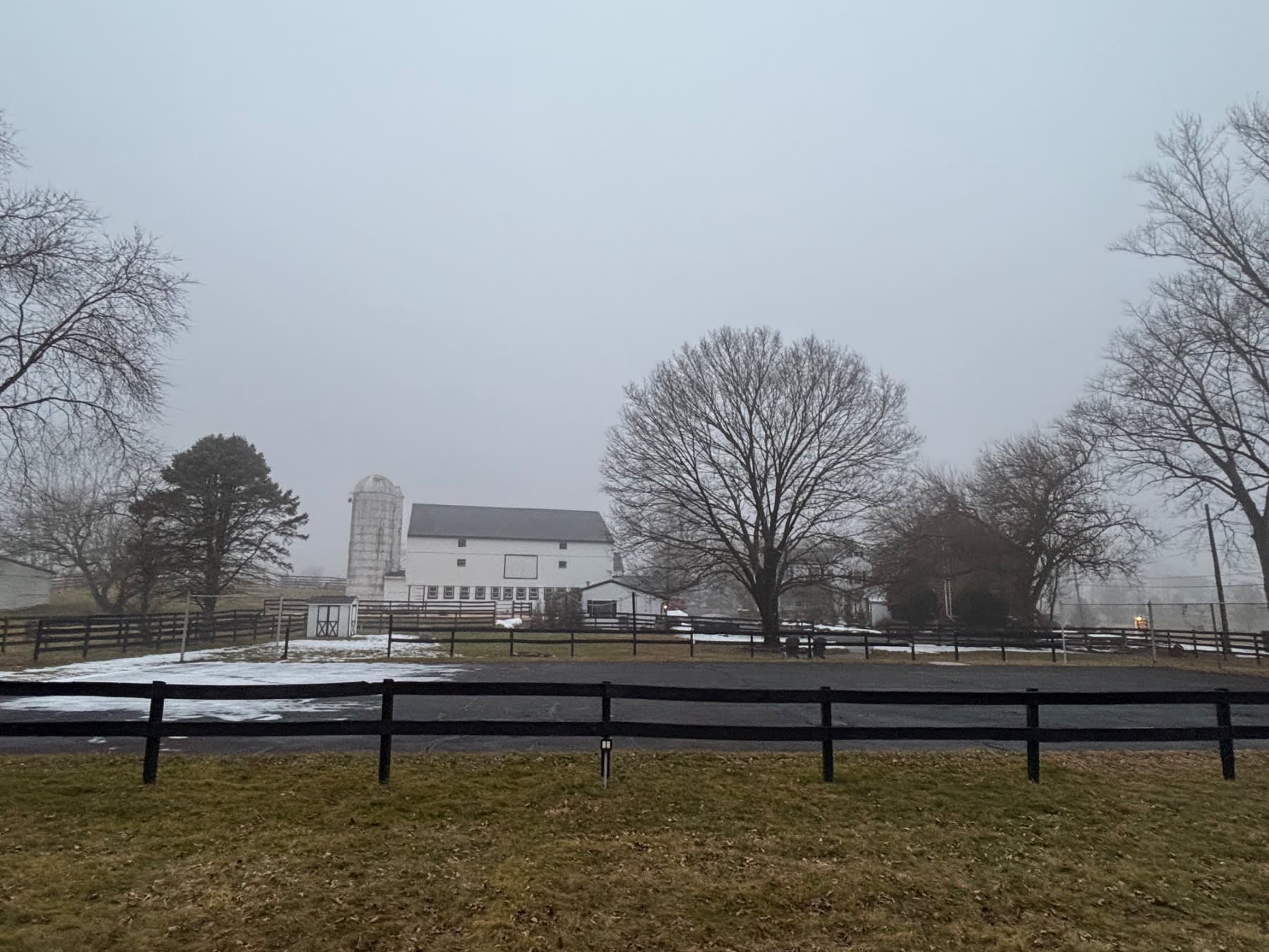 A white barn with silo stands behind wooden fences in a misty rural landscape with bare winter trees.