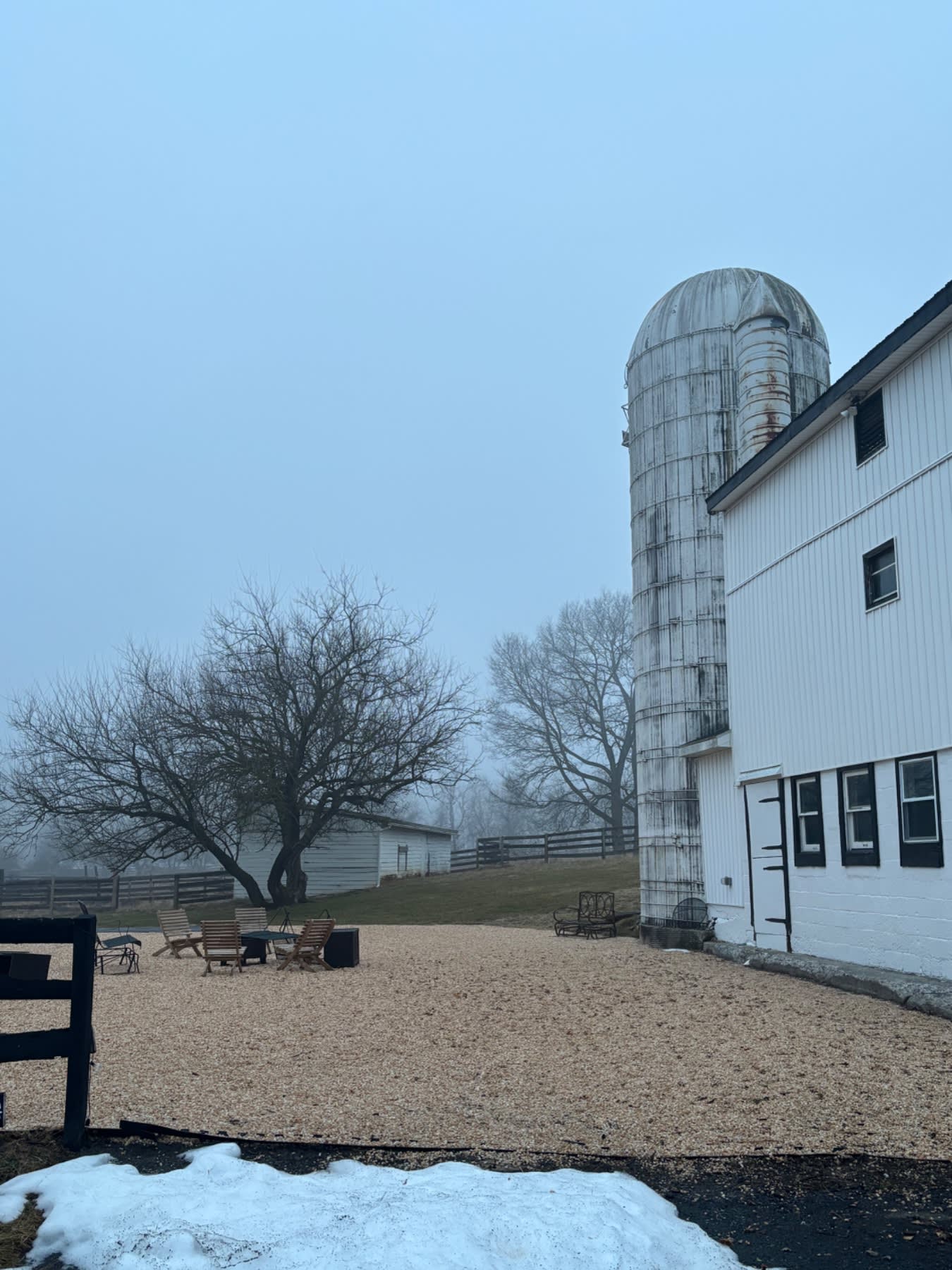 # Alt Text
A white farmhouse with a tall metal silo stands beside a gravel courtyard with bare trees and wooden fencing under a clear blue sky.