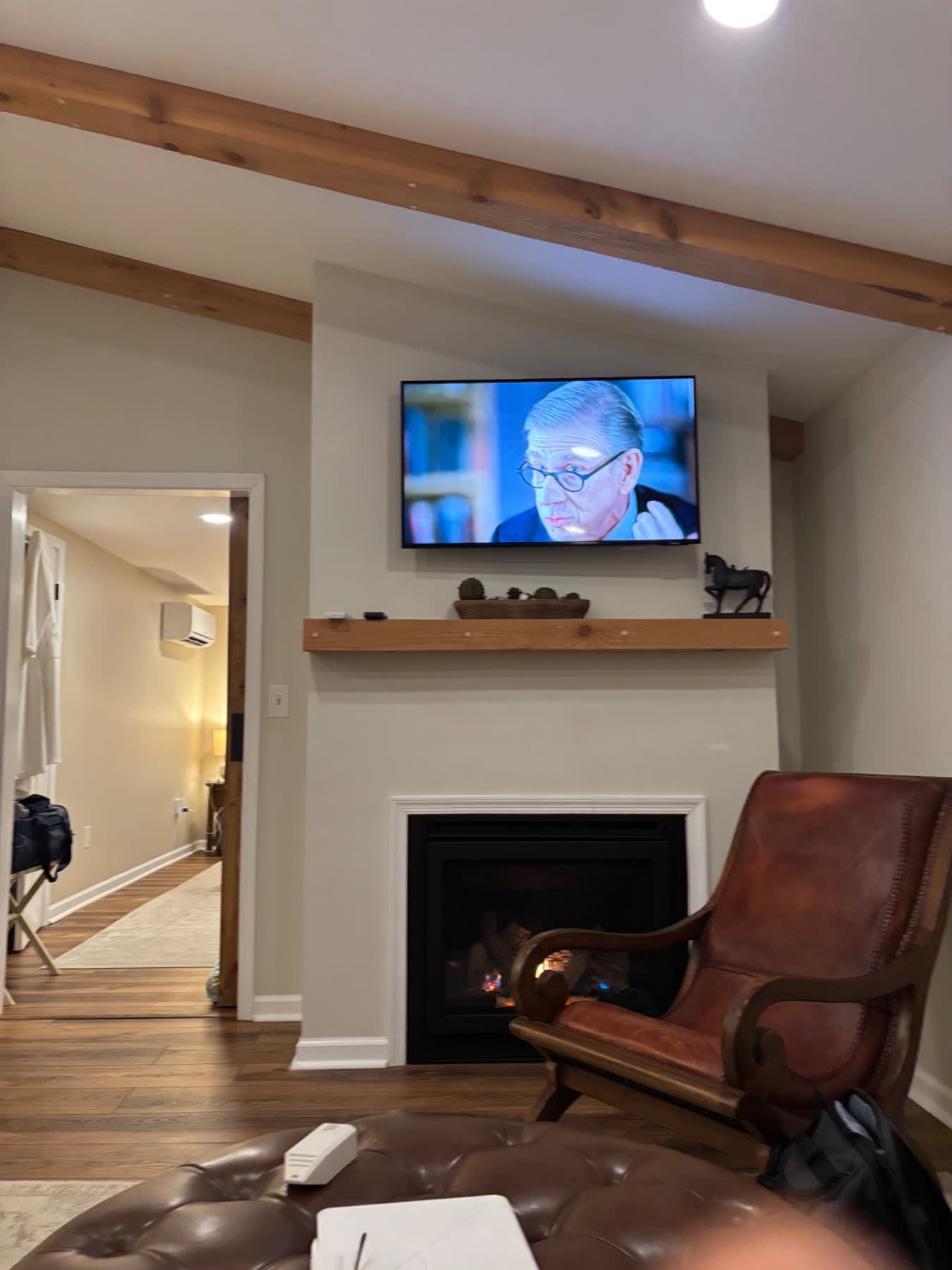 A modern living room with a fireplace, wooden beam ceiling, mounted TV, leather armchair, and open doorway to another room.