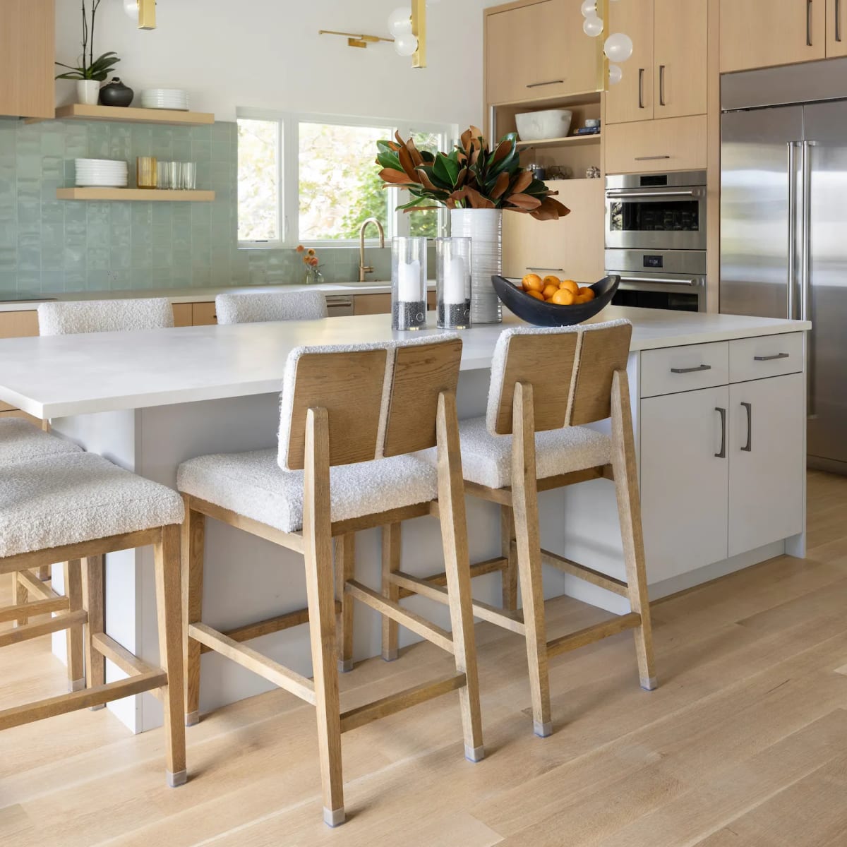 White oak kitchen with a seafoam green tile backsplash, brass globe pendants and boucle counter stools at a white quartz island