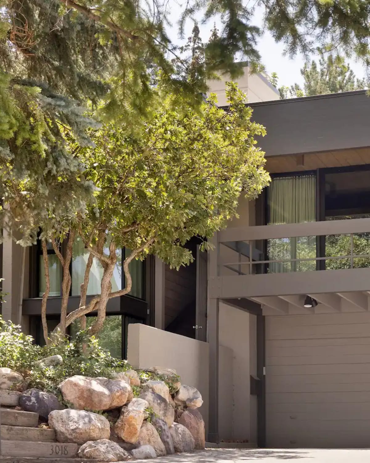 Midcentury modern home exterior with a low-pitched roof and clerestory windows, set among mature trees and boulder landscaping. Organic Midcentury, Salt Lake City, UT