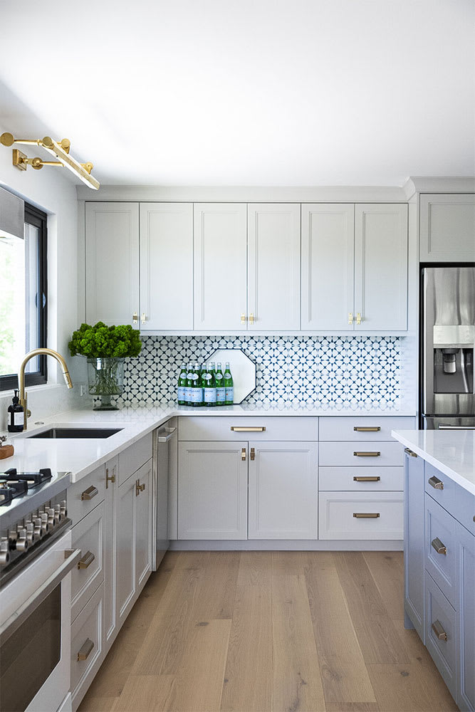 Pale grey kitchen with a blue and white patterned tile backsplash, brass swing-arm sconce and green hydrangeas