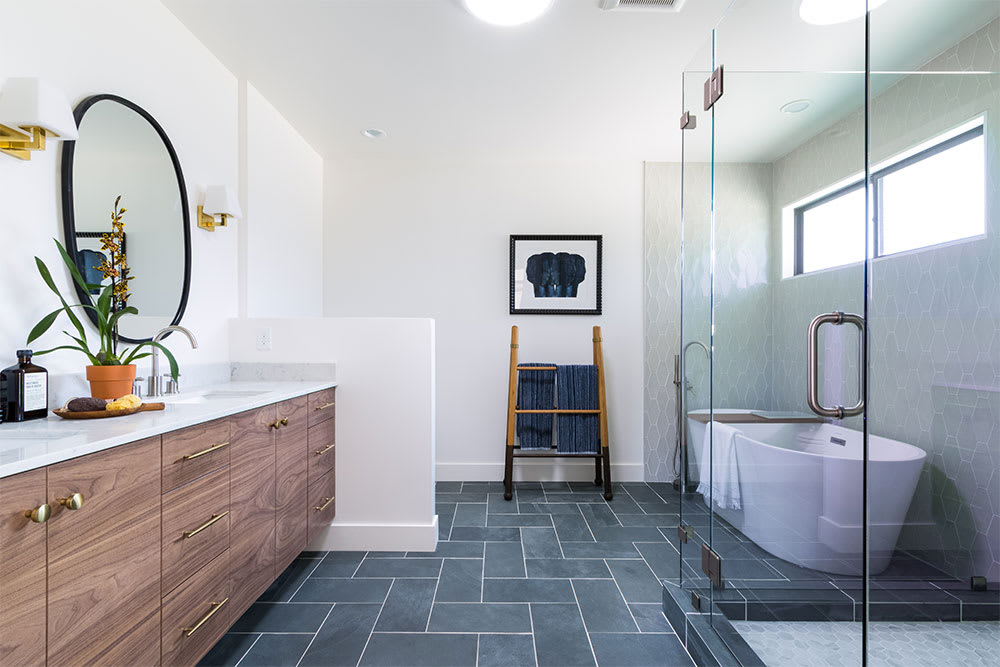 Bathroom with a walnut double vanity, oval black mirror and freestanding tub above slate grey floor tile