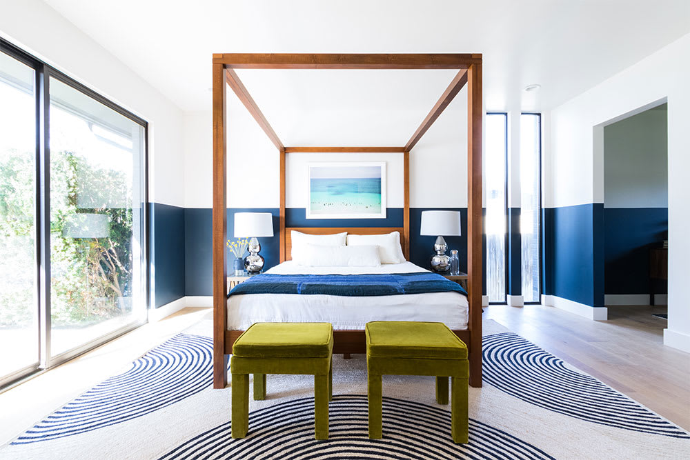 Bedroom with a wooden four-poster bed against a two-tone navy wall, chartreuse velvet stools and a graphic monochrome rug