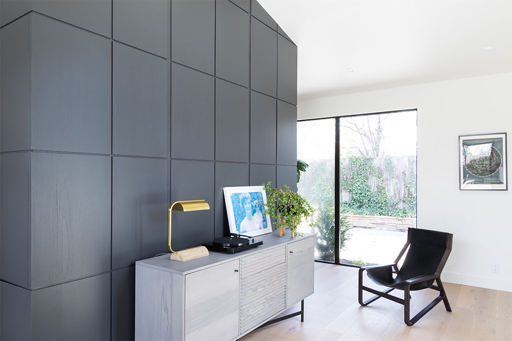 Charcoal panelled wall beside a pale wood credenza with a record player and brass lamp, and a black leather chair