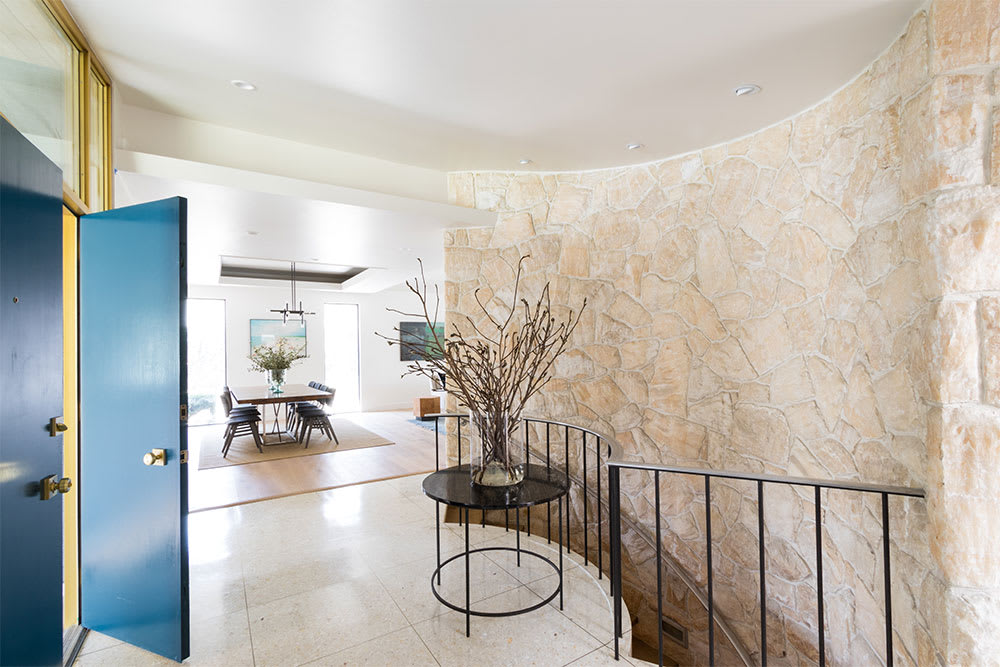 Entry with a blue front door, curved cut-stone wall and black metal stair railing beside a round table of branches