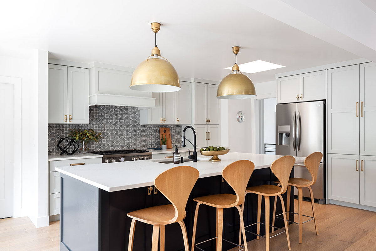 Black kitchen island with bentwood barstools beneath brass dome pendants, against white cabinetry and a grey tile backsplash