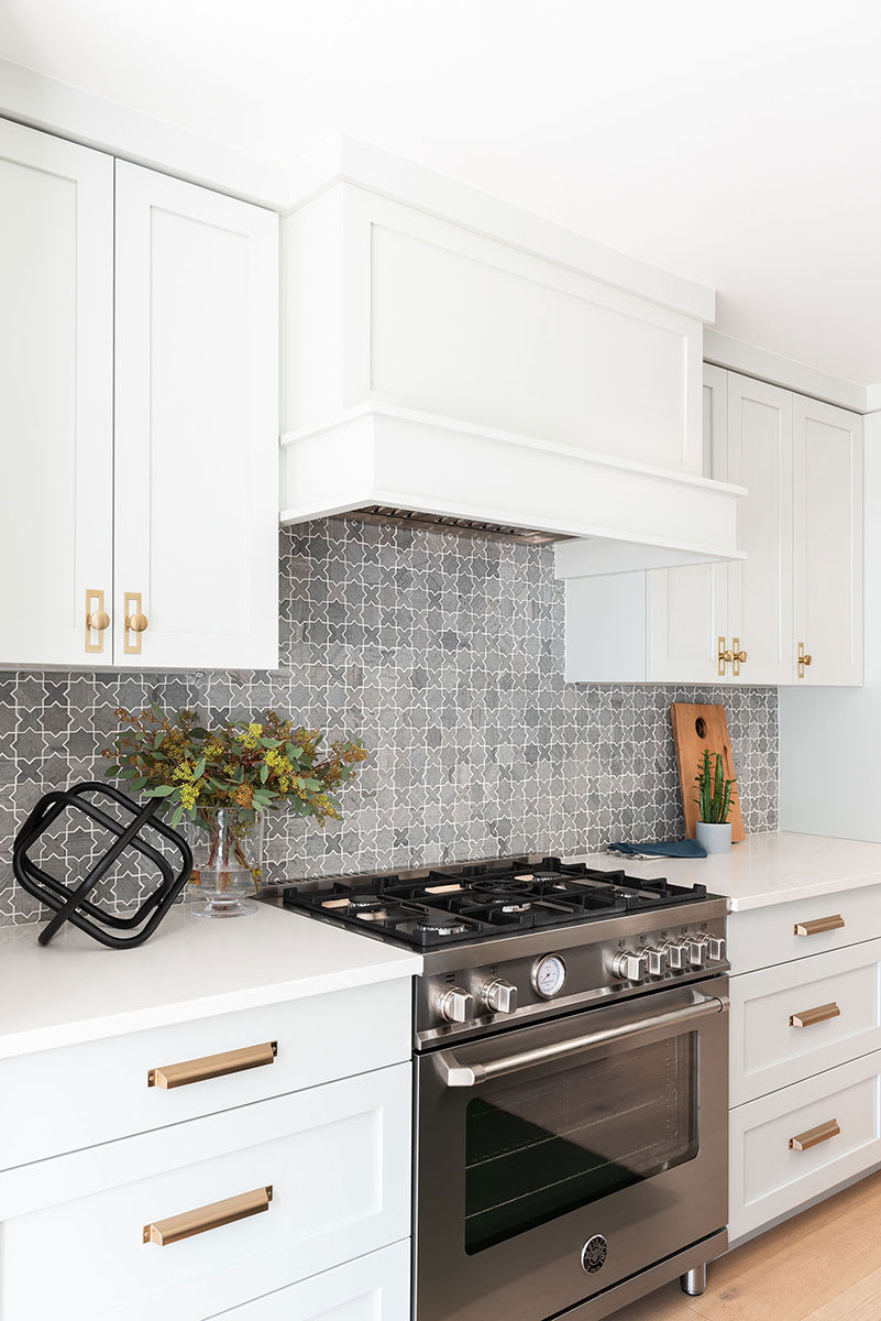 White shaker kitchen with a panelled hood, grey patterned tile backsplash, stainless range and brass hardware