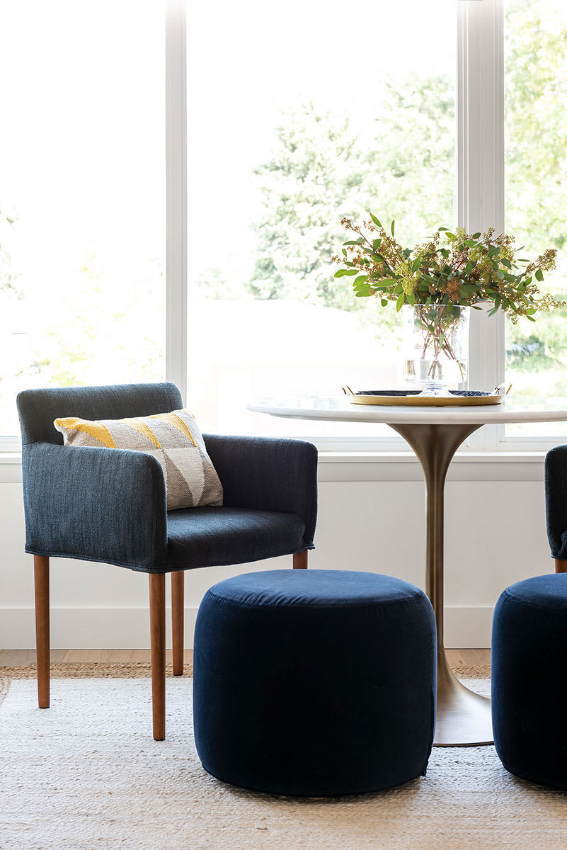 Window nook with a blue upholstered armchair, navy velvet ottomans and a round pedestal table of flowers