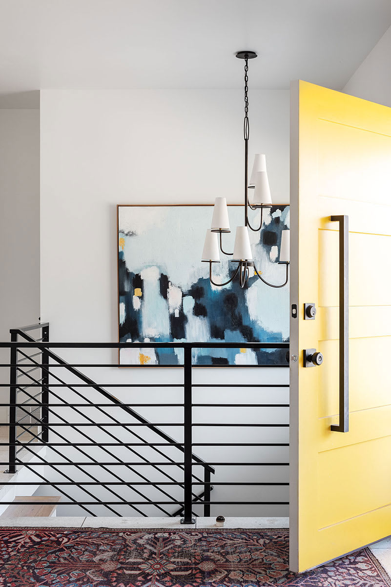 Entry with a bright yellow front door, black horizontal stair railing and a blue abstract painting under a candle chandelier