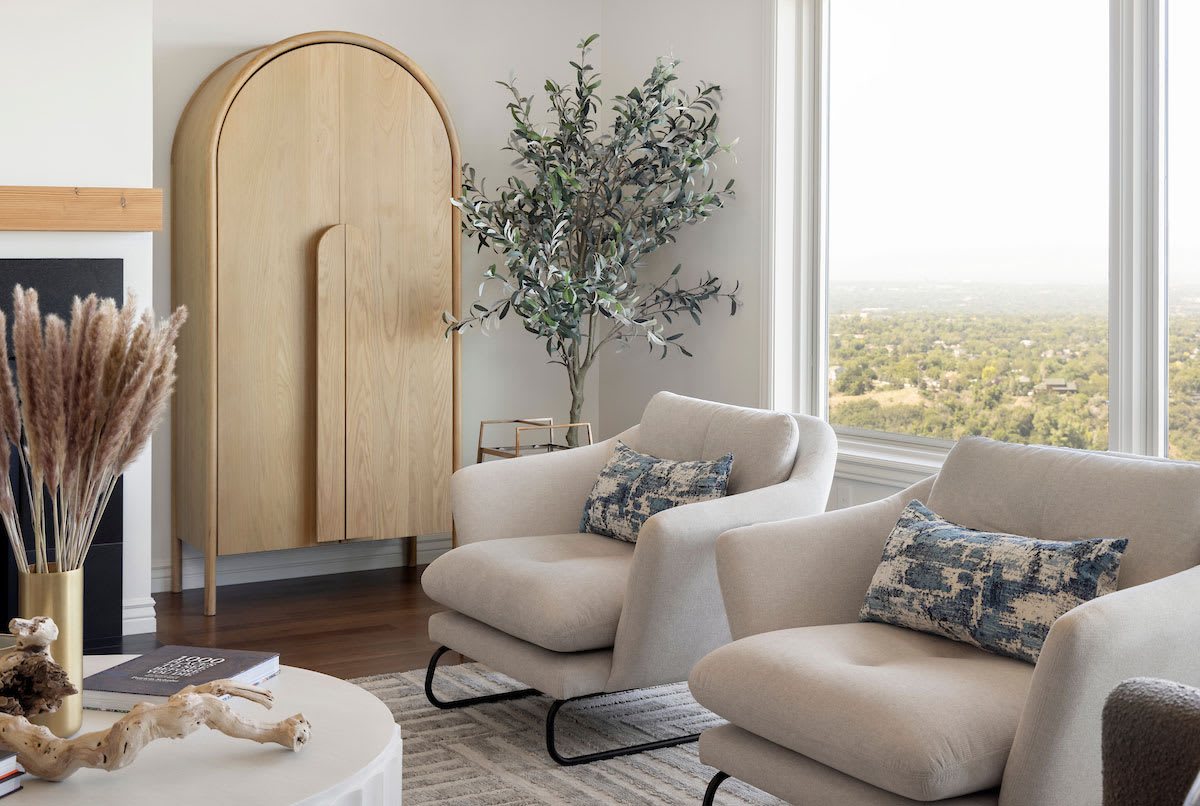 Two cream lounge chairs with blue cushions beside a tall arched oak cabinet and an olive tree, with a valley view through the corner windows