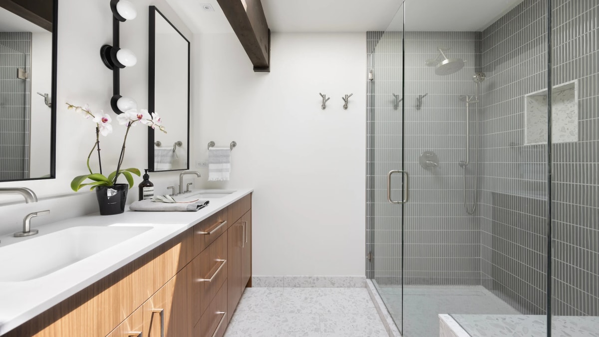 Bathroom with a walnut double vanity, grey stacked-tile walk-in shower and an orchid on the counter