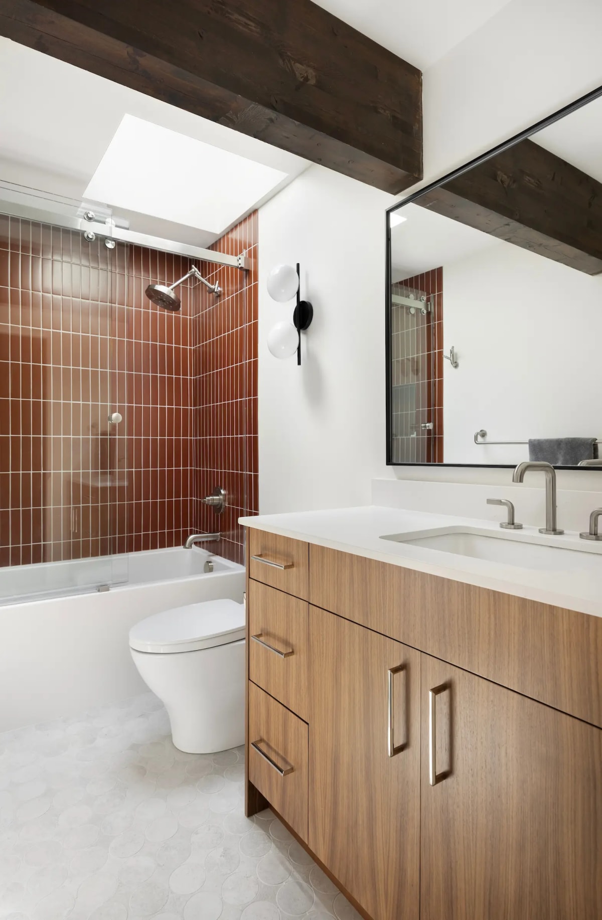 Bathroom with a rust terracotta tiled shower-tub, walnut vanity and skylight above, with a black wall sconce