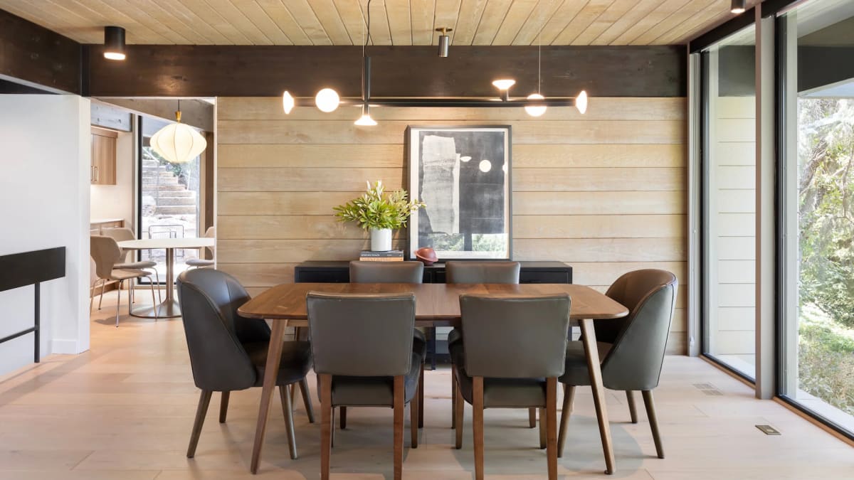 Walnut dining table with grey leather chairs against a pale wood plank wall, lit by a linear pendant