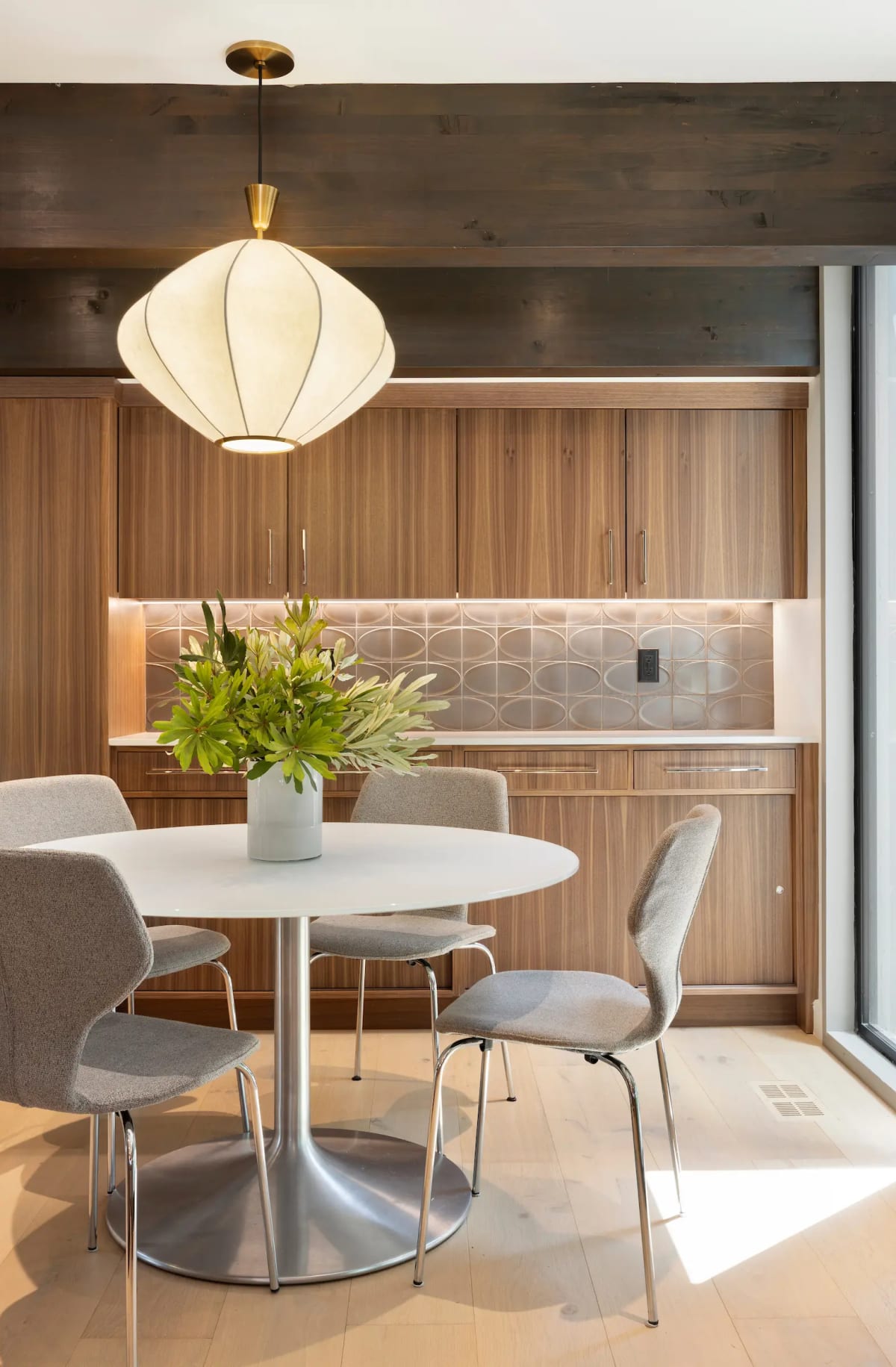 Breakfast nook with a round white pedestal table and grey chairs against walnut cabinetry and a patterned tile backsplash
