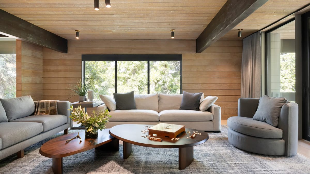 Wood-clad living room with exposed dark beams, two cream sofas, grey swivel chairs and sculptural walnut coffee tables