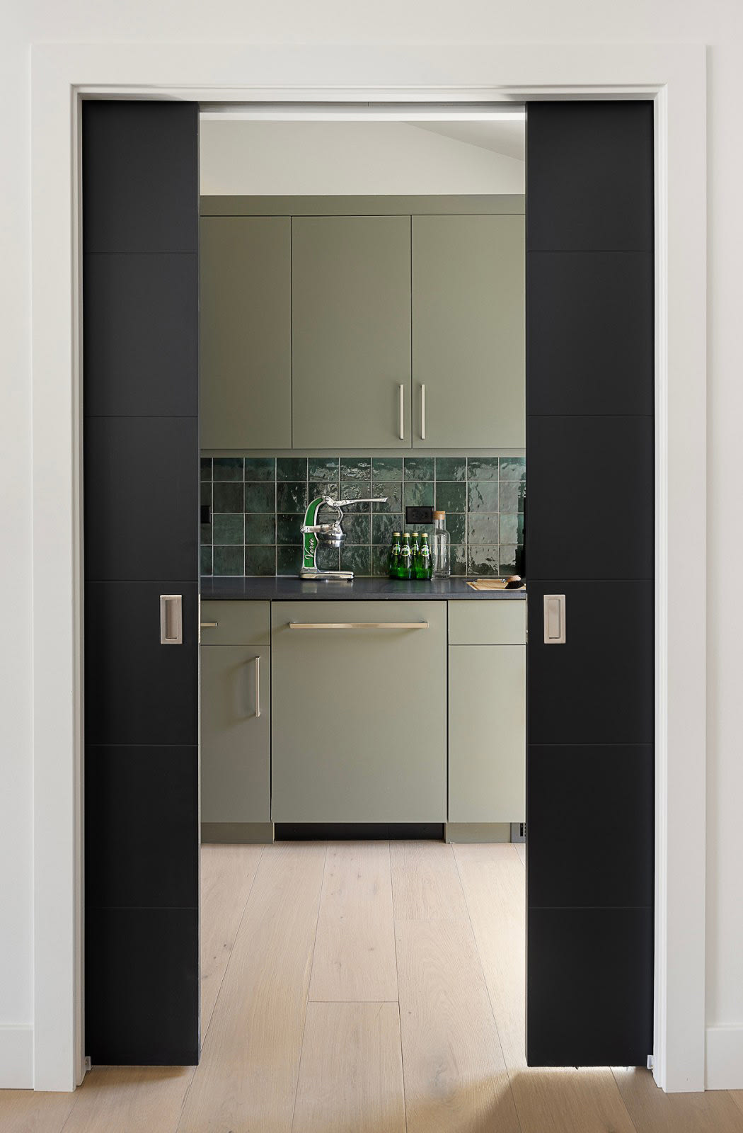 Black pocket doors opened to a sage green butler's pantry with a green glazed tile backsplash