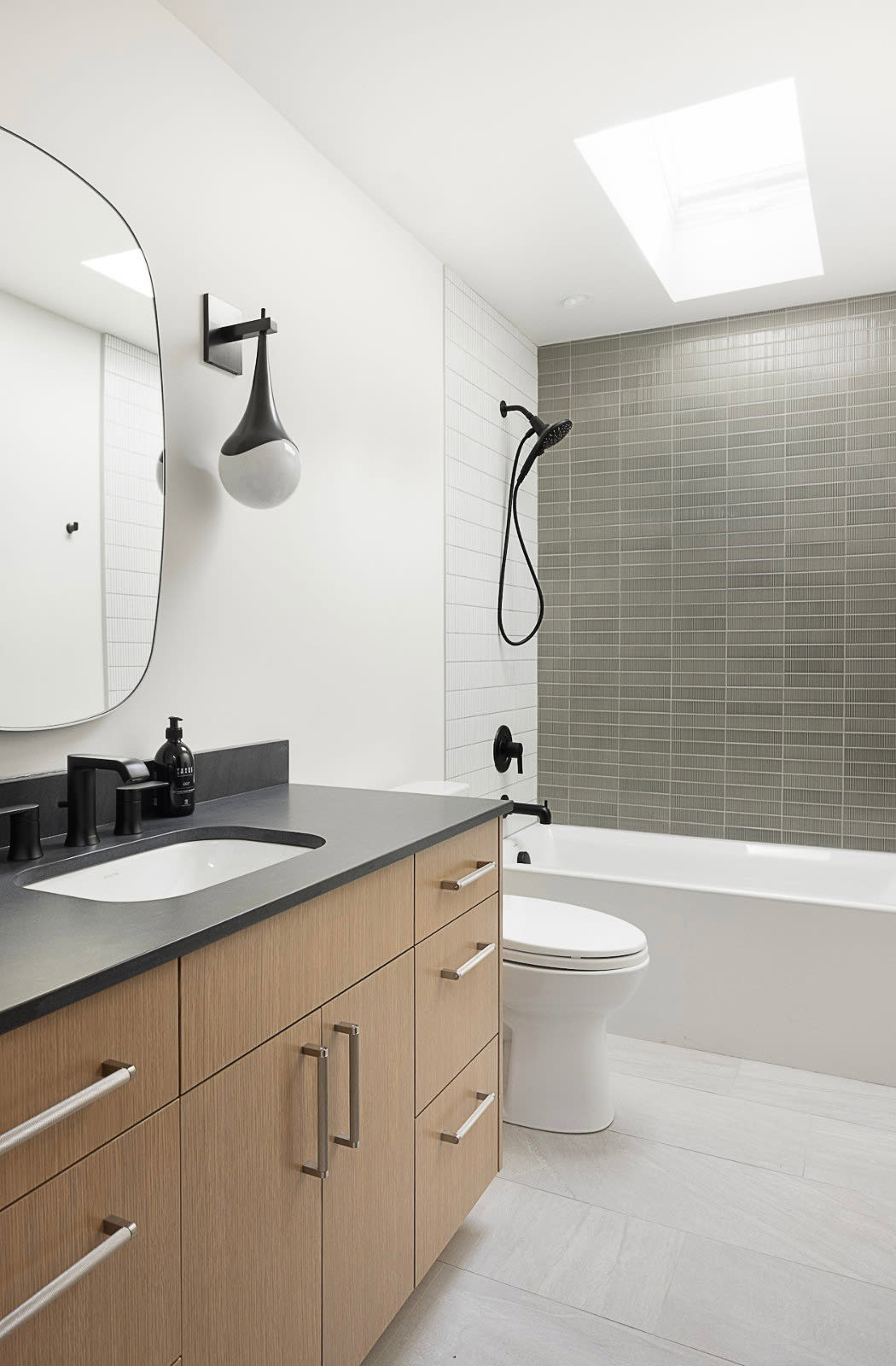 Bathroom with a white oak vanity and black stone counter, arched mirror and skylight above an olive tiled tub surround