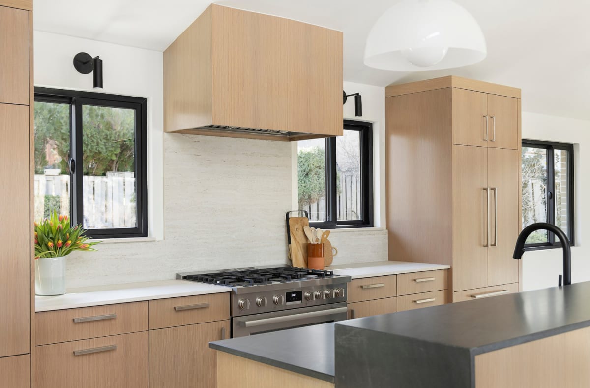 White oak kitchen with a matching hood over a stainless range, stone slab backsplash and black-framed windows