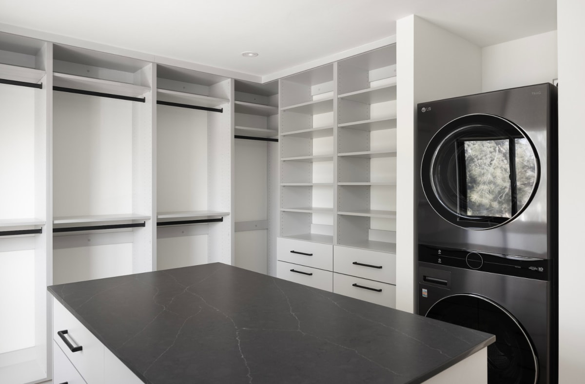 Combined laundry and dressing room with open white shelving, a black stone island and a stacked washer and dryer