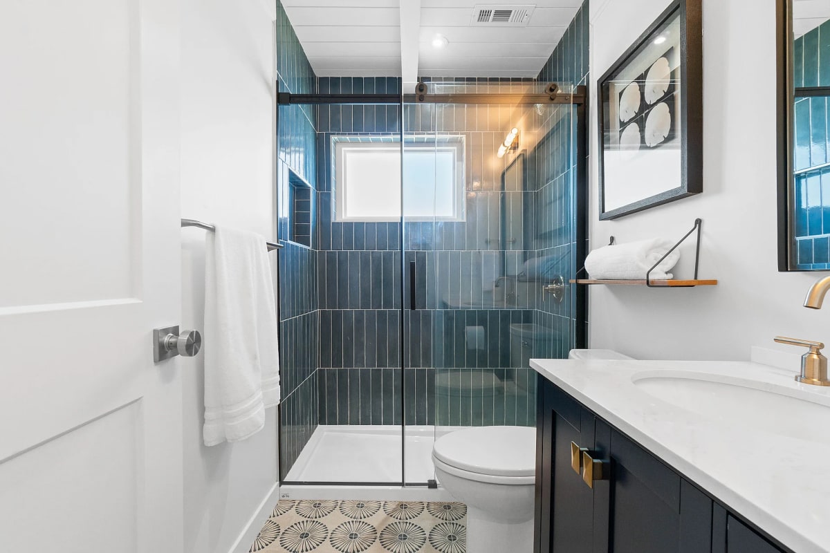 Bathroom with a teal stacked-tile walk-in shower, navy vanity with brass fixtures and patterned floor tile