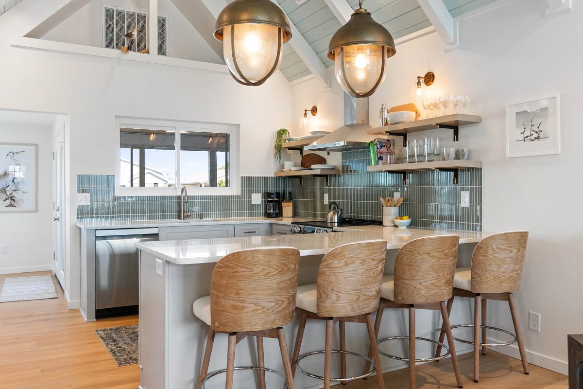Kitchen peninsula with a blue-grey tile backsplash, white quartz counter, four oak barstools and industrial pendant lights