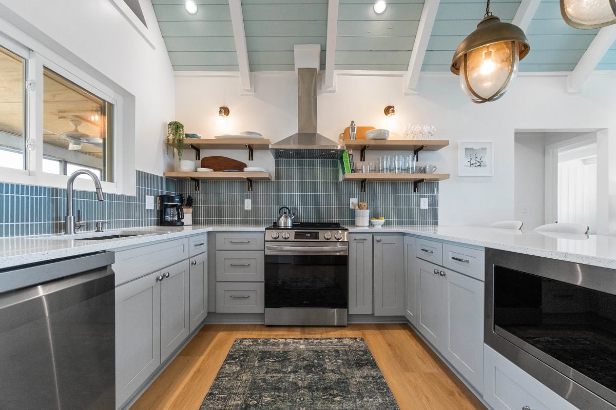 Grey shaker kitchen with a blue vertical tile backsplash, stainless range and open wood shelving under a seafoam plank ceiling
