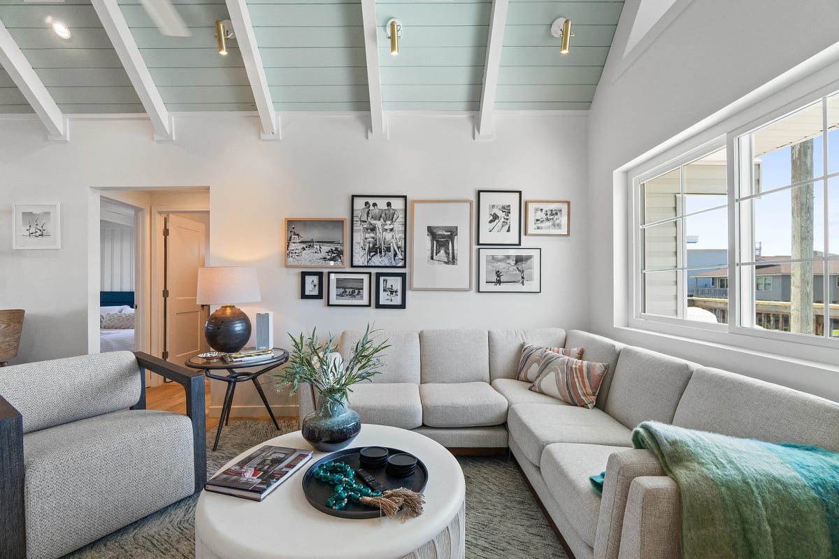 Cream sectional beneath a gallery wall of black and white beach photographs, under an exposed seafoam beamed ceiling