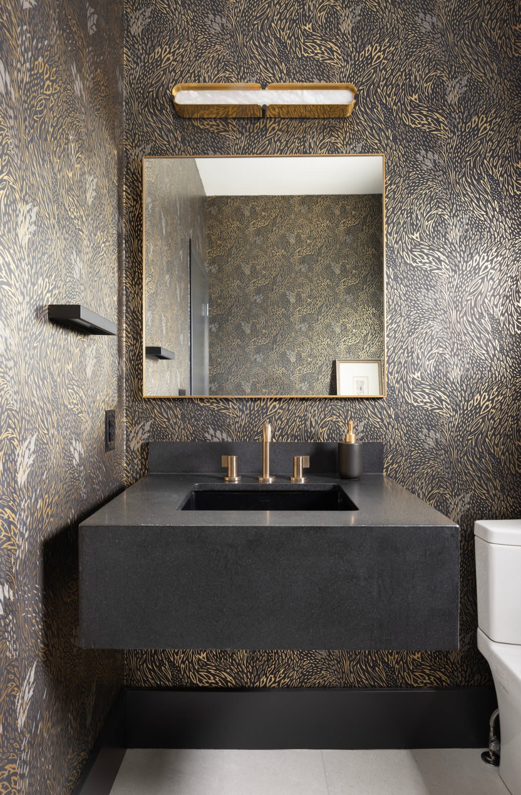Powder room with metallic gold patterned wallpaper, a floating dark stone vanity and brass fixtures under a marble sconce