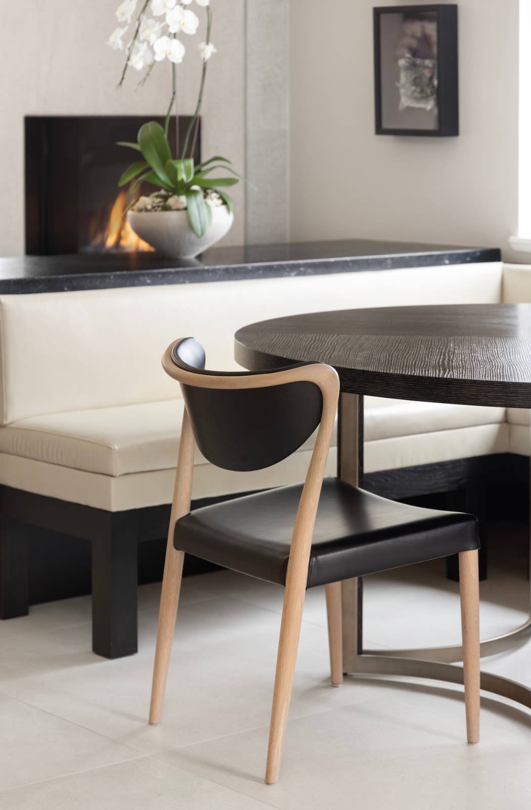 Built-in cream banquette and dark round table with a blond wood chair, beside a linear gas fireplace and orchid