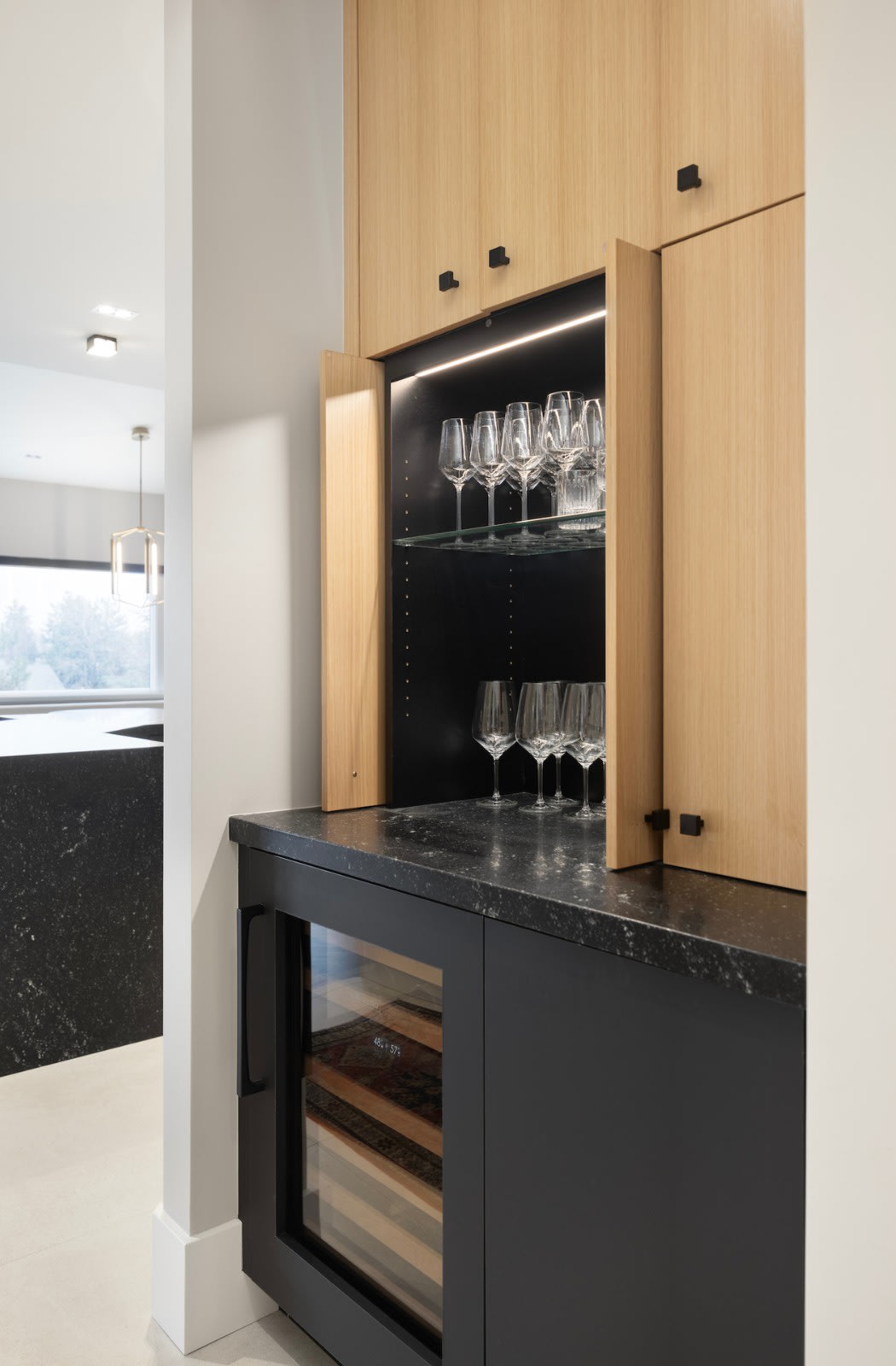 Built-in bar with white oak doors, lit glass shelving and a black stone counter above a wine refrigerator