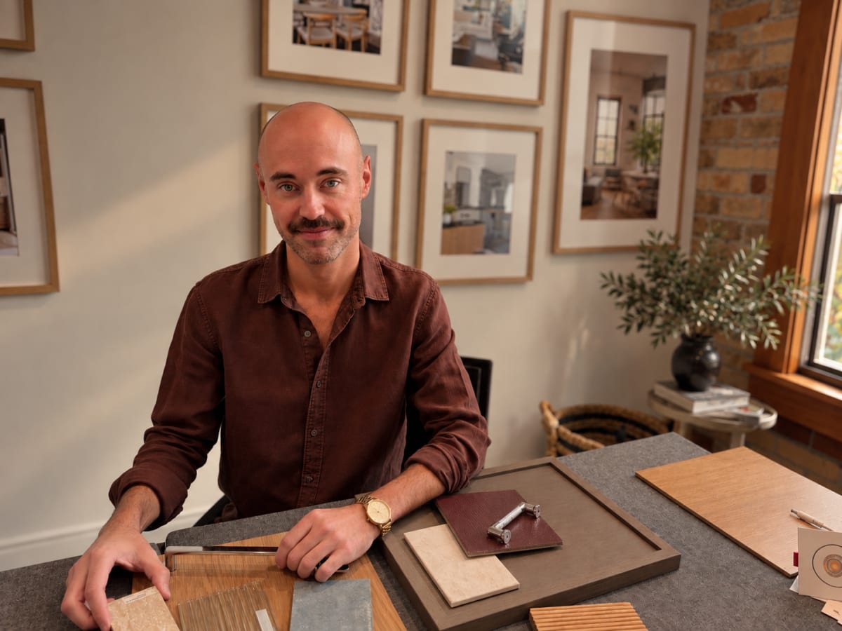 Alex Kirby Bowman at a desk in the studio, with finish samples and framed interiors photography behind him