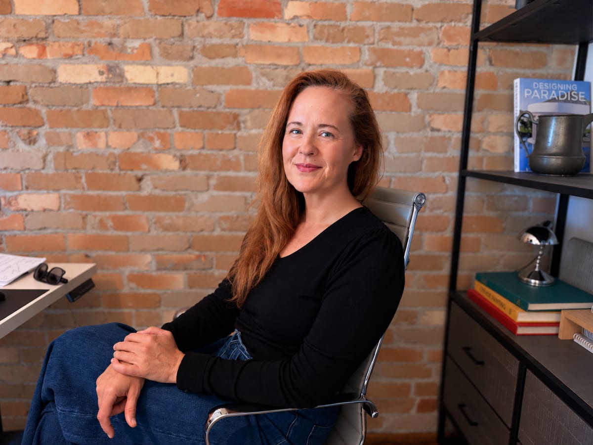 Leane Jensen seated in the studio against an exposed brick wall, beside shelves of design books