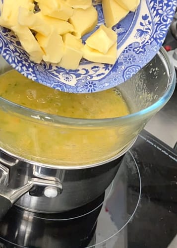 Small pieces of butter being added to the whisked mixture in a bowl over a saucepan half filled with hot water