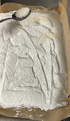 Dried coconut being spread on top of meringue spread evenly into the prepared Swiss roll tin.