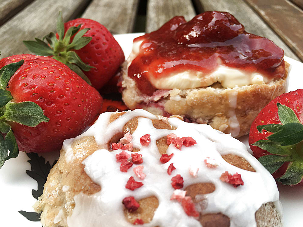 Strawberry and Rose scone cut in half on a plate, spread with clotted cream and strawberry jam and with whole fresh strawberries.