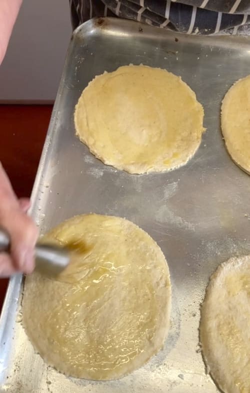 Four round bases on a greased baking tray being brushed them with a little olive oil.