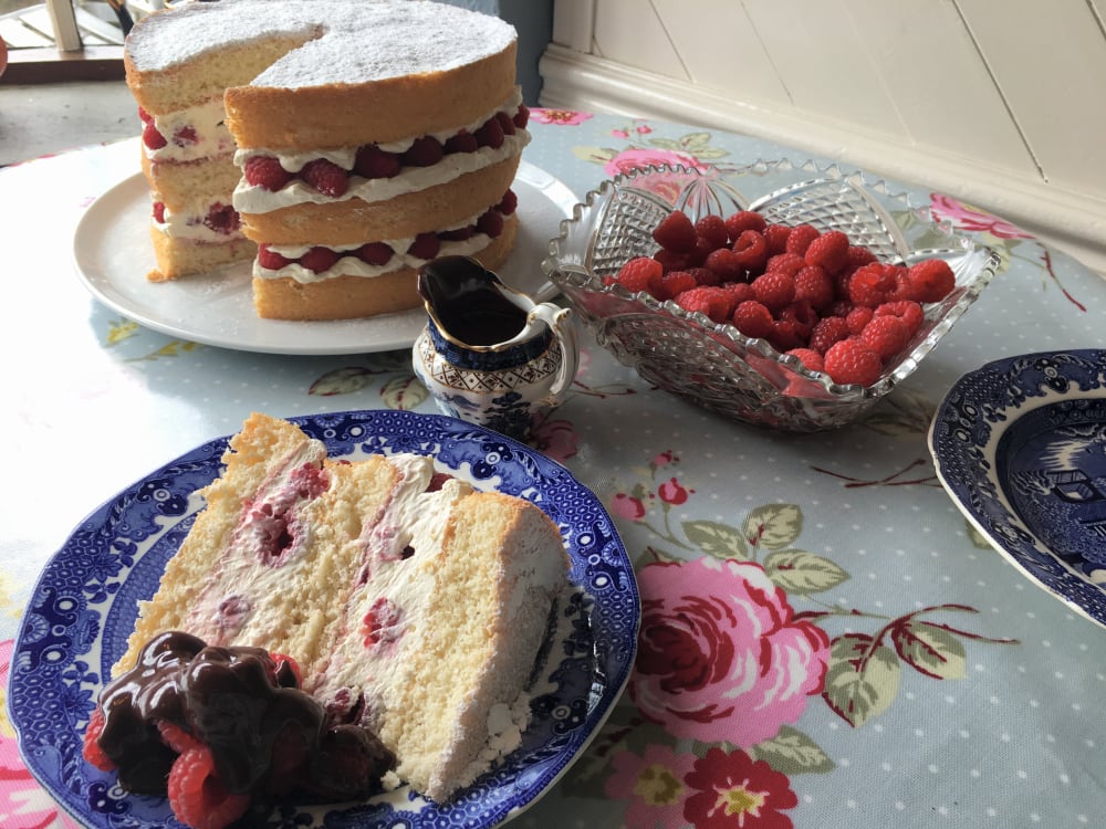 Slice of raspberry cream sponge on a plate with a bowl of fresh raspberries, jug of chocolate sauce and the full sponge in the background