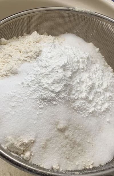 Flour, sugar and baking powder being sieved through a colander