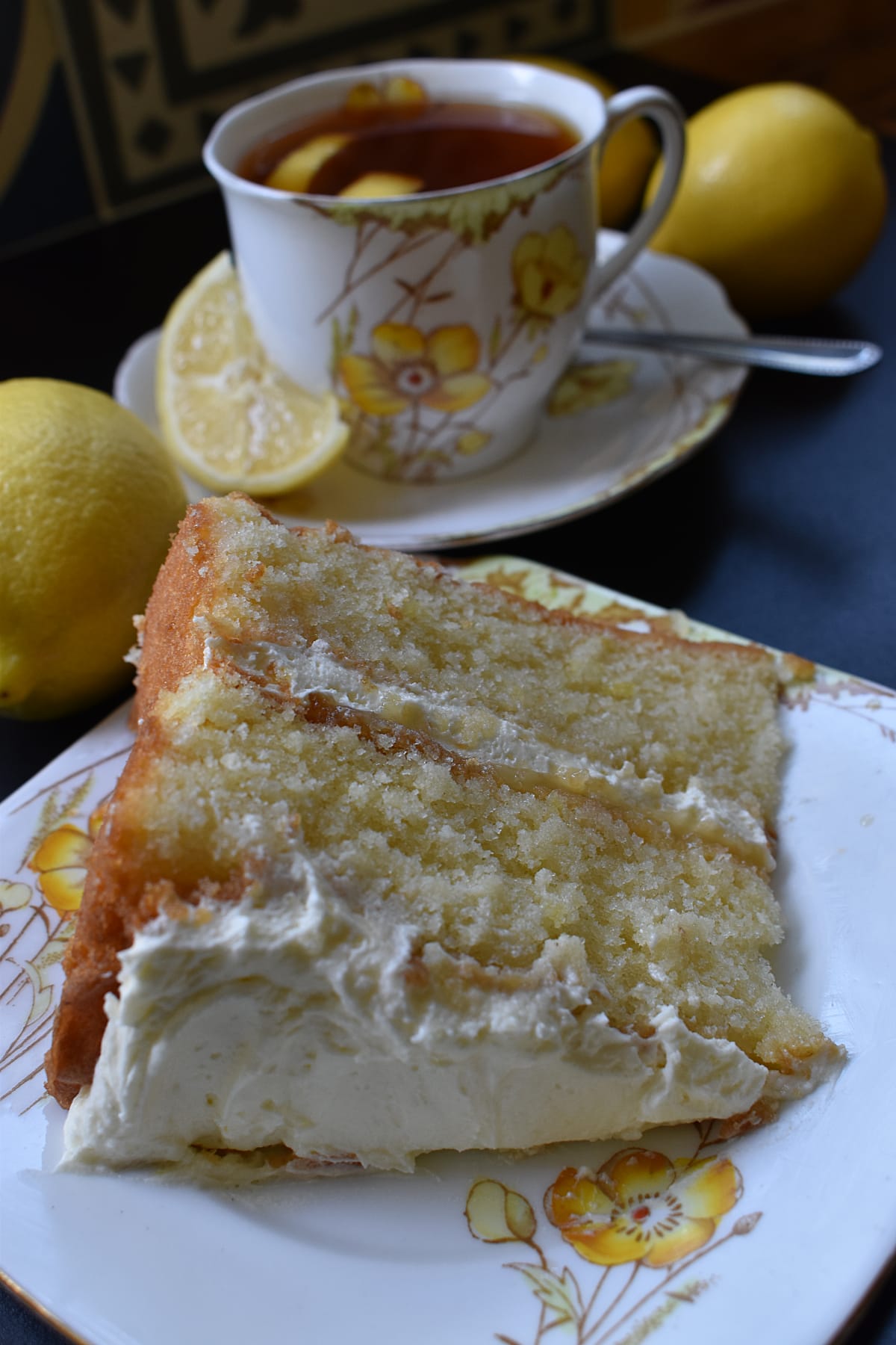 Slice of Lemon Cake on a plate with cup of tea and lemons in the background.