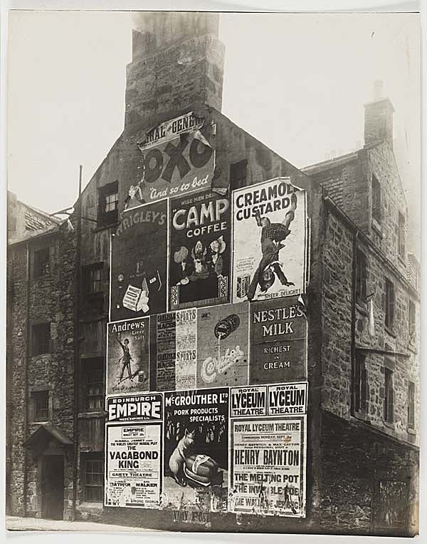 Photograph of a three storey tenement building in Edinburgh, Scotland covered in eleven large poster advertisements. The posters cover one entire side of the building and are for a variety of different products. On the right there is a washing line with some laundry hanging from it. Some of the posters are simply text eg. Edinburgh Empire, while others include images eg. Camp Coffee.