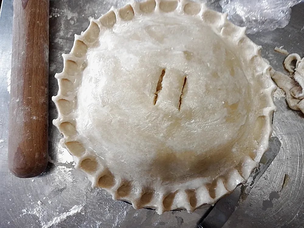 Uncooked whole apple pie on work surface alongside wooden rolling pin